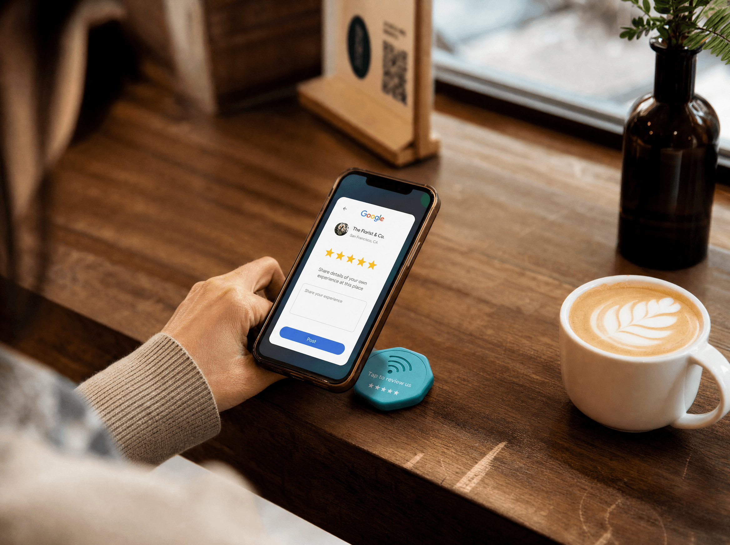 Customer at café table tapping phone to a teal Tapora NFC badge — phone shows 5-star Google Review prompt